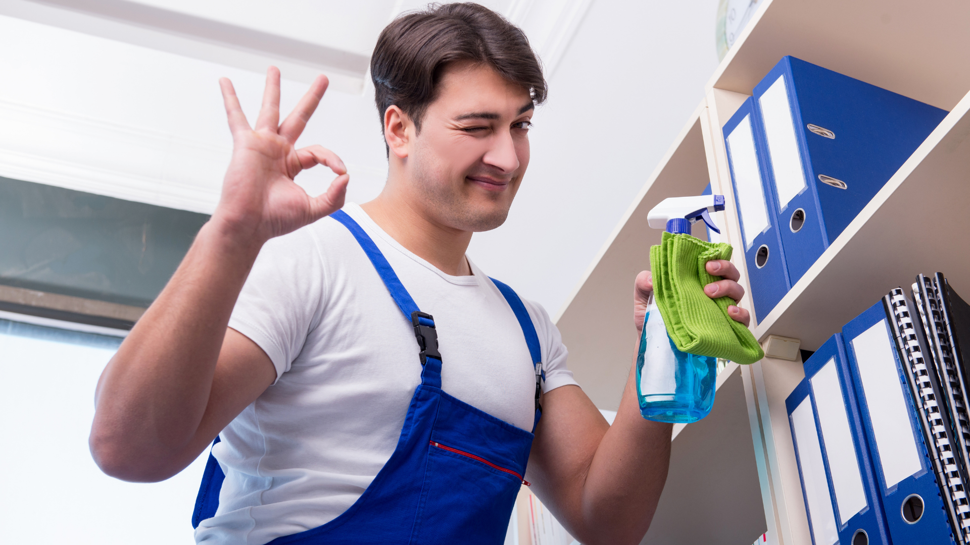 Add a subheading (3) A person in blue overalls winks and makes an "OK" hand sign while holding a spray bottle and cleaning cloth in front of a shelf with binders and files.