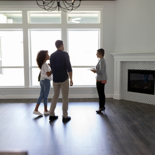 A couple stands in an empty, bright living room with large windows, talking to a real estate agent holding a tablet next to a fireplace.