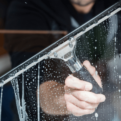 A person uses a squeegee to clean a soapy, wet glass window, with water and soap suds visible on the glass surface.