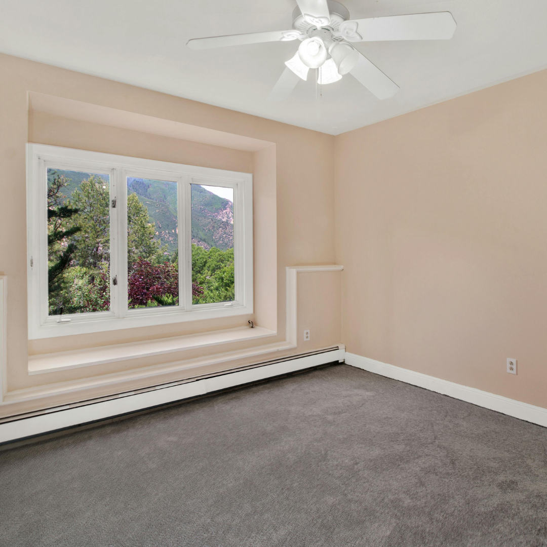 Empty beige room with gray carpet, white trim, a ceiling fan, and a large window with mountain and tree views. Built-in shelves are on either side of the window seat area.