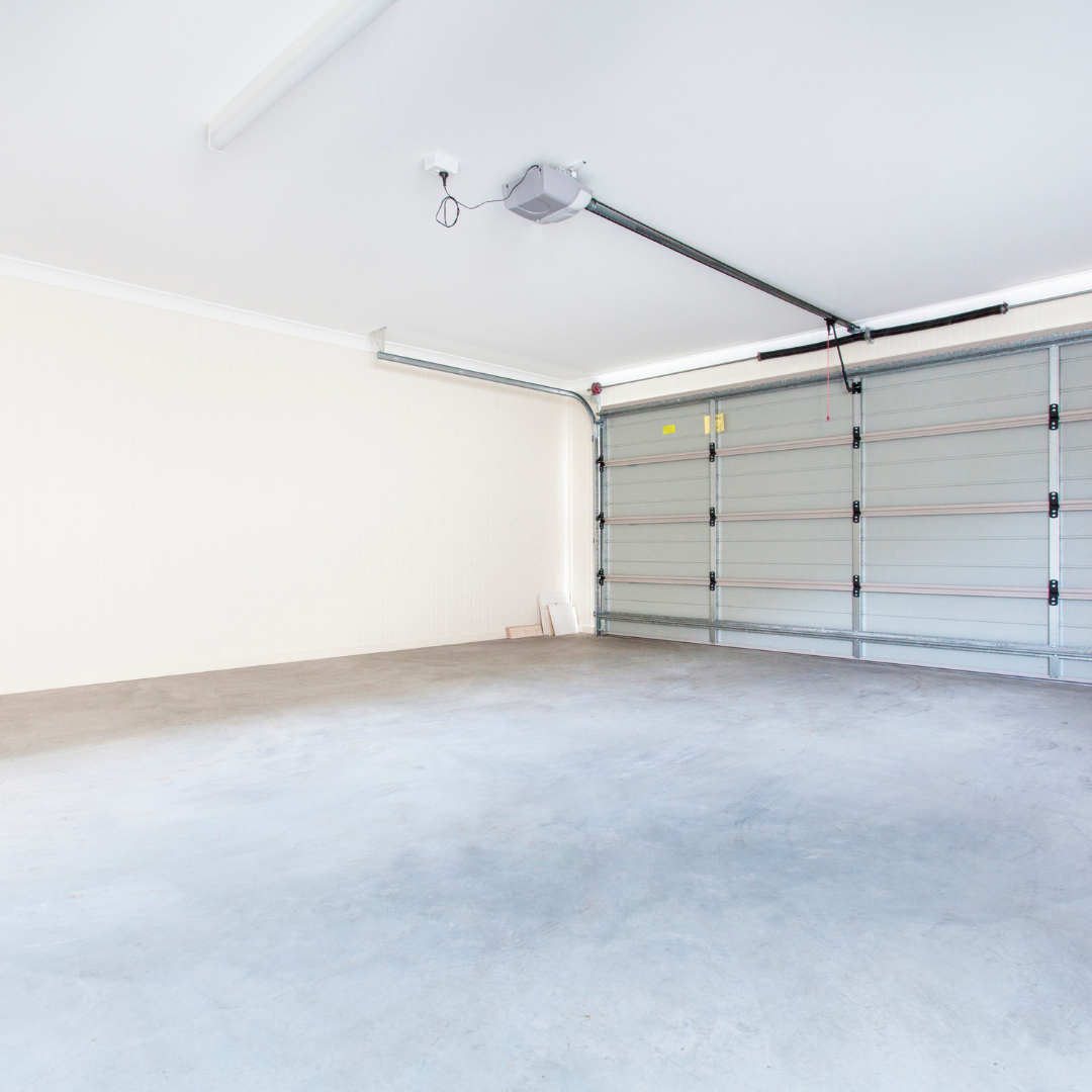 A clean, empty garage with a smooth concrete floor, off-white walls, and a large closed sectional garage door. Some objects are stacked against the far wall. The space is brightly lit.