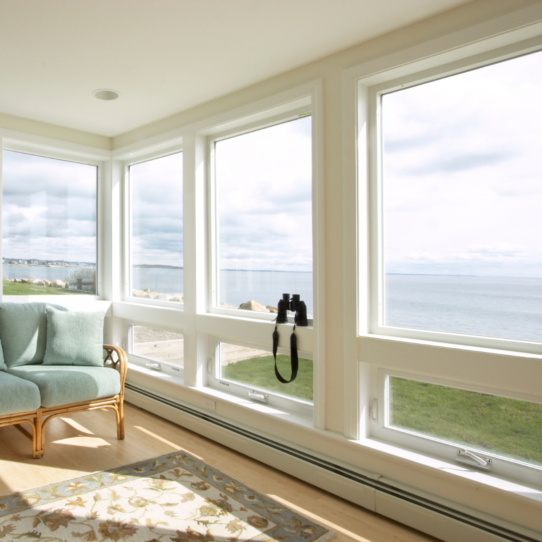 A bright sunroom with large windows overlooks a calm ocean and cloudy sky. A wicker chair with light blue cushions and a floral rug sit by the window, along with black binoculars resting on the window sill.
