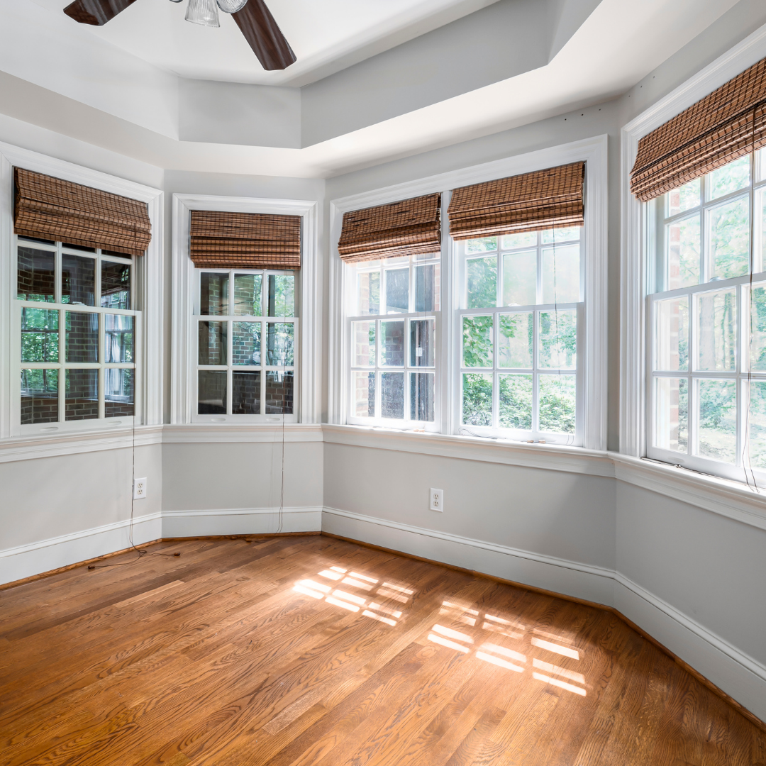 A bright, empty room with hardwood floors, light gray walls, and large windows with brown woven blinds. Sunlight streams through the windows, creating patterns on the floor. Ceiling fan blades are visible above.