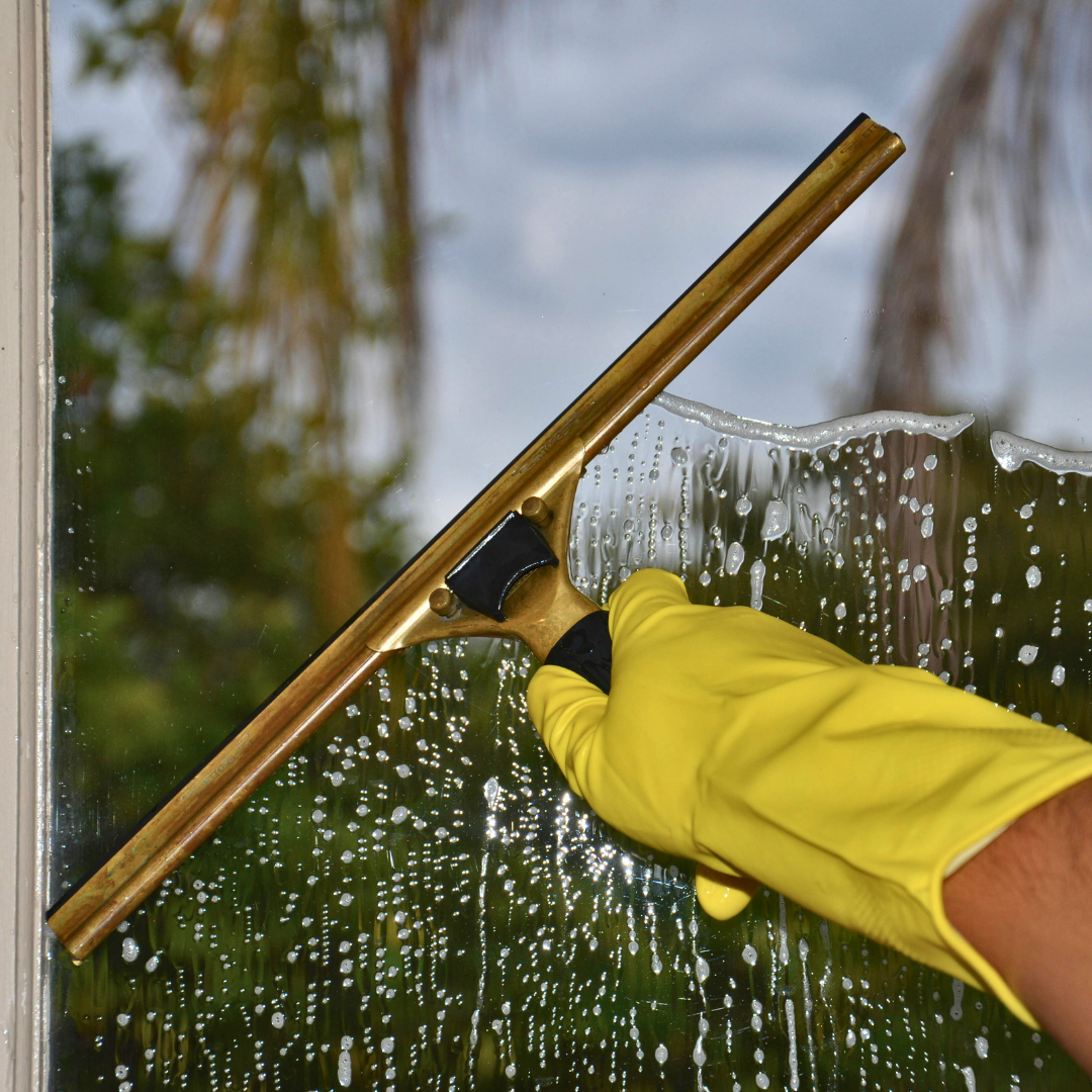 A person wearing a yellow rubber glove uses a brass squeegee to clean soapy water off a window, with trees and a cloudy sky visible in the background.