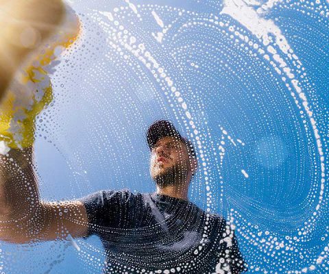 A man wearing a dark t-shirt and cap is seen from below while cleaning a glass surface with soapy water against a bright blue sky.