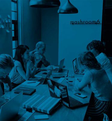 Five people sit at a table working on laptops in a modern office with notebooks, water bottles, and phones. The wall has "mashroom6" written on it. The image has a blue color filter.